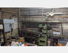 Werkstatt mit einer Bohrmaschine und einer grünen Metallfräsmaschine. Umgeben von Regalen mit Metallstangen, Werkzeugen, Kunststoffbehältern und verschiedenen Arbeitsmaterialien, in einem rustikalen Raum mit unverputzten Wänden.