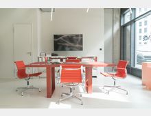 Moderner Besprechungsraum mit einem roten Tisch und vier passenden roten Bürostühlen auf hellweißem Boden. An der Wand hängt ein großer Flachbildfernseher, daneben eine Glastür, und rechts befindet sich eine Fensterfront mit Blick auf eine Straße.