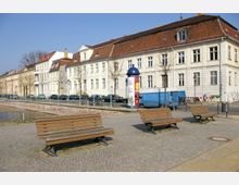 Straßenszene mit Altbauhäusern im Hintergrund, gepflastertem Gehweg und mehreren Holzbänken im Vordergrund. Im Hintergrund verläuft ein niedriger Kanal, und daneben stehen geparkte Autos sowie ein blauer Transporter.