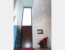 Heller Eingangsbereich mit hoher Decke, einer großen dunklen Holztür mit bodentiefem Fenster, einem kleinen Fenster rechts und einer leuchtend roten Sitzbank. Links steht ein blauer Schrank, der Boden ist aus Holz und ein roter Teppich liegt vor der Tür.