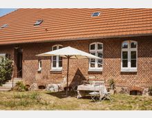 Backsteinfassade eines ländlichen Gebäudes mit weißen Fensterrahmen und einem roten Ziegeldach, davor eine kleine Terrasse mit einem weißen Sonnenschirm, einem gedeckten Tisch und zwei Stühlen auf einer grasbewachsenen Fläche.