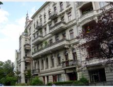 Mehrgeschossiges Wohngebäude im historischen Stil mit verzierten Fassaden, Balkonen und hohen Fenstern. Im Vordergrund sind Bäume und ein kleiner Gartenbereich zu sehen, die Umgebung wirkt urban.