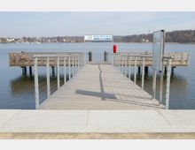 Holzsteg mit Metallgeländern, der in einen ruhigen See führt, mit zwei seitlichen Aussichtsplattformen. Im Hintergrund ist ein Ufer mit Bäumen und Gebäuden sowie ein Schild mit Informationen zu Fährdiensten zu sehen.