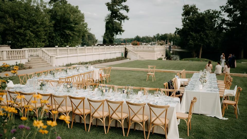 Lange, festlich gedeckte Tafeln mit weißen Tischdecken, Glasgeschirr und Holzstühlen stehen auf einer Rasenfläche im Freien. Im Hintergrund verläuft eine weiße Balustrade entlang einer Terrasse mit Bäumen und einem Wasserlauf daneben.