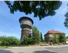 Ein historischer Wasserturm mit einer gemauerten Basis und einer Holzkonstruktion im oberen Teil, umgeben von Bäumen und Sträuchern. Rechts daneben steht ein zweistöckiges Haus mit roten Dachziegeln und einem eingezäunten Vorgarten.