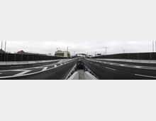 Eine leere, mehrspurige Autobahn mit schwarzen Schallschutzwänden auf beiden Seiten. Im Hintergrund sind Gebäude und Verkehrsschilder zu sehen, der Himmel ist bewölkt.