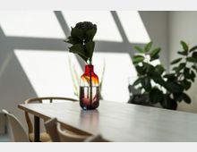 Ein schlichter, heller Raum mit einem Holztisch, auf dem eine rote Glasvase mit einer Pflanze steht. Im Hintergrund sind grüne Zimmerpflanzen und geometrische Schattenmuster an der Wand erkennbar.