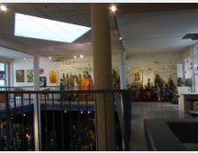 Ein zweistöckiger Ausstellungsraum mit einer Galerieebene, die durch Geländer abgegrenzt ist. Die obere Etage zeigt Kunstwerke an den Wänden und eine Sammlung von Figuren auf Podesten, während die untere Etage teilweise sichtbar ist und durch eine helle Deckenöffnung beleuchtet wird.