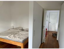 Ein Schlafzimmer mit einem Holzbett auf der linken Seite, bedeckt mit zerknitterter, beigefarbener Bettwäsche, und ein angrenzender Flur mit Holzboden, der zu einer geöffneten weißen Tür führt.