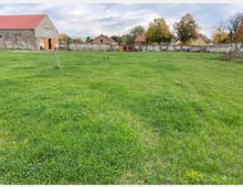 Weite Wiese mit grünem Gras, umgeben von einer niedrigen Mauer. Im Hintergrund stehen mehrere Gebäude mit roten Dächern, darunter ein großes steinernes Gebäude auf der linken Seite und Bäume mit Herbstlaub.