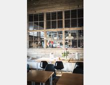 Ein gemütlicher Innenraum eines Cafés mit Holztischen und schwarzen Stühlen. Im Hintergrund ist eine Wand aus grobem Holz mit großen Fenstern zu sehen, die Einblick in einen weiteren Raum mit dekorativen Bildern und Gegenständen bietet.