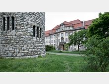 Ein historisches Gebäude mit heller Fassade, dekorativen Steinverzierungen und roten Ziegeldächern steht hinter einem Rasen und Bäumen. Links im Bild ist der Teil eines Rundbaus aus Stein zu sehen, mit kleinen Fenstern verbunden durch dicke Mauern.