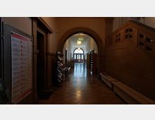 Flur in einem historischen Gebäude mit gewölbtem Steinbogen, gefliesten Boden und Holzbänken entlang der Wände. Links sind ein Schaukasten und Bücherregale zu sehen, während am Ende des Flurs eine große Glastür mit seitlichen Fenstern durch Tageslicht beleuchtet wird.