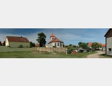 Dorfansicht mit Fachwerkkirche im Zentrum, umgeben von Wohnhäusern mit roten Dächern und einem kleinen Gartenbereich. Eine Backsteinmauer und ein unbefestigter Weg verlaufen im Vordergrund.