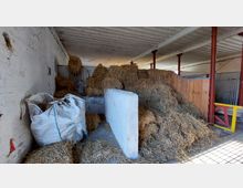 Innenraum eines landwirtschaftlichen Stalls mit großen Mengen an gestapeltem Stroh. Im Vordergrund befinden sich ein weißer großer Sack und eine Betonabtrennung; rote Metallpfeiler und Holzelemente strukturieren den Raum.