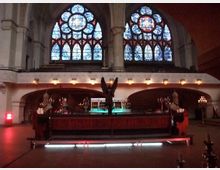Innenraum einer Kirche mit großen, farbigen Glasfenstern im gotischen Stil. Im Vordergrund steht ein aufwendig gestalteter Altar mit Kerzen, darüber thront ein schwarzer Adler, und der Raum ist dezent beleuchtet.