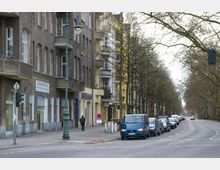 Straßenszene in einer städtischen Umgebung mit mehrstöckigen Wohnhäusern, teils grauem und teils gelbem Putz, darüber hinaus geparkten Autos entlang der Straße und kahlen Bäumen am Straßenrand. Ein Fußgänger geht den Gehweg entlang, und ein Laternenmast steht in der Nähe.