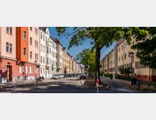 Straßenansicht in einem Wohnviertel mit mehrstöckigen Häusern in warmen Farben wie Orange, Gelb und Beige, teilweise mit Schrägdächern. Auf beiden Seiten der Straße sind geparkte Autos, Bäume spenden Schatten, und ein Gehweg mit Straßenschildern ist erkennbar.