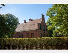 Backsteinhaus mit geschwungener Giebelfront und mehreren Schornsteinen auf dem roten Ziegeldach, umgeben von Hecken, Bäumen und einem Holzzaun, im Hintergrund blauer Himmel.