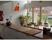 Helle Küche und Essbereich mit großen bodentiefen Fenstern, die Blick auf einen Garten und Backsteinfassaden bieten. Ein langer Esstisch aus Holz mit Stühlen steht neben einer Küchenzeile mit weißen Schränken und einer Holzarbeitsfläche, umgeben von Pflanzen und dekorativen Elementen.