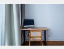 Ein minimalistischer Arbeitsplatz mit einem Holzschreibtisch, auf dem ein Computer mit Bildschirm, Tastatur und Maus steht. Vor dem Tisch steht ein Holzstuhl mit Rattangeflecht, daneben helle Vorhänge und eine weiße Wand.