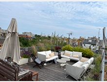 Dachterrasse mit Holzboden, modernen weißen Sitzmöbeln und einem großen Sonnenschirm, umgeben von Pflanzen in Kübeln; im Hintergrund eine städtische Wohngegend mit mehrstöckigen Gebäuden und grünem Baumbestand.