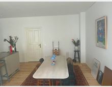 Wohnzimmer mit hellem Holzboden, einem langen Esstisch mit Stühlen und einer roten Teppichläufer. Im Hintergrund eine weiße Tür, links eine Anrichte mit Dekorationen und rechts ein Rahmenbild an der weißen Wand.