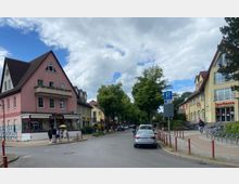 Straßenszene in einer kleinen Stadt mit mehreren Gebäuden; links ein rosa Wohnhaus mit Geschäft im Erdgeschoss, rechts ein gelbes Gebäude mit Sparkassen-Filiale. Die Straße ist von Bäumen gesäumt, mit parkenden Autos und Fußgängern auf dem Gehweg.