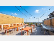 Rooftop terrace with gray paver flooring, orange rectangular tables, and white metal chairs beneath strings of hanging lights. Wooden slat walls frame the space, with planters along the edge and a wide city skyline view in the distance.