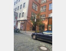 Eine städtische Gasse mit Kopfsteinpflaster, flankiert von einem Gebäude aus roten Backsteinen und einer weißen Fassade mit Fenstern. Im Vordergrund steht ein geparktes Auto, während Fahrräder an einem Zaun links angelehnt sind; ein Baum und ein verglaster Eingang sind im Hintergrund sichtbar.