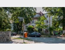 Straßenecke mit einem zweistöckigen Wohnhaus im klassischen Stil, umgeben von Bäumen und einem Metallzaun. Im Vordergrund befinden sich Straßenschilder der "Malvenstraße" und "Limonenstraße", ein orangefarbener Mülleimer, geparkte Autos und ein Fahrradfahrer auf einer gepflasterten Straße.