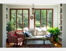 Helles Wohnzimmer mit großen, holzgerahmten Sprossenfenstern zur Gartenseite, vor denen ein beiges Sofa, ein gestreifter Sessel und ein niedriger Couchtisch auf einem gemusterten Teppich stehen. Seitliche Regale, mehrere Zimmerpflanzen und eine hängende Deckenlampe ergänzen den Raum.