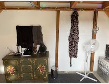 Innenraum eines Raumes mit einer schlicht gestrichenen weißen Wand und sichtbaren Holzbalken. Im Vordergrund steht eine grün lackierte Kommode mit Blumenmustern, darauf liegen verschiedene Alltagsgegenstände. Rechts hängen ein Bademantel mit Giraffenmuster und zwei Handtücher; daneben steht ein weißer Standventilator auf einem grauen Teppich. Entlang der Decke ist eine Lichterkette angebracht.