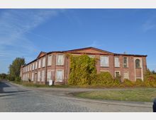 Backstein-Gebäude mit zwei Stockwerken, dessen Fenster größtenteils durch Bretter verschlossen sind. Die Fassade ist teilweise mit Efeu bewachsen, und das Gebäude steht an einer gepflasterten Straße in einer ländlichen Umgebung.