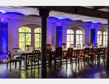 Industrieller Veranstaltungsraum mit einer langen, gedeckten Tafel und gemischten Stühlen vor großen, bogenförmigen Fenstern. Die Beleuchtung ist in blau gehalten, und die Wände zeigen eine rohbelassene Betonstruktur.