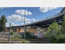 Eisenbahnbrücke über Bahngleise, umgeben von mit Graffiti besprühten Wänden und Vegetation. Im Hintergrund sind ein gelbes Gebäude, Metallkonstruktionen und ein teils bewölkter Himmel sichtbar.