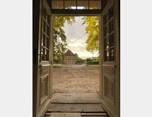 Blick durch geöffnete Holztüren auf einen Hof mit sandigem Boden, umgeben von Bäumen. Im Hintergrund steht ein Fachwerkhaus, darüber ein teils bewölkter Himmel in warmem Licht.