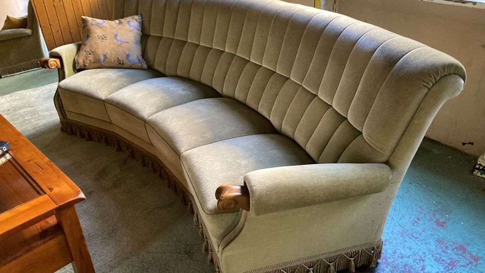 Gebogener Wohnraum mit großem, gepolstertem Samtsofa in hellem Grau mit Fransenbesatz und Holzarmlehnen, davor ein Holztisch. Im Hintergrund stehen hohe Fenster und eine holzvertäfelte Wand, der Boden ist teils mit Teppich, teils mit abgenutztem Anstrich versehen.