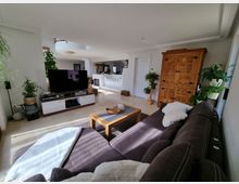 Modernes Wohnzimmer mit heller Einrichtung; im Vordergrund eine braune Stoffcouch, daneben ein kleiner Holztisch. Im Hintergrund stehen ein Flachbildfernseher auf einem weißen TV-Schrank, ein großer Holzschrank und mehrere grüne Zimmerpflanzen. Helle Wände und Fliesenboden verleihen dem Raum eine helle und offene Atmosphäre.