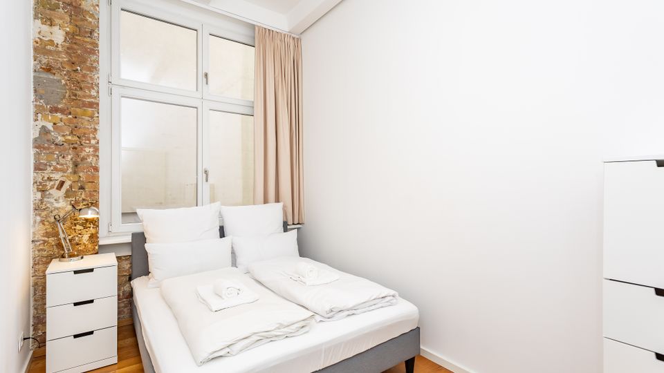 Helles, minimalistisches Schlafzimmer mit Doppelbett und weißer Bettwäsche, neben einem kleinen Nachttisch. Große Fenster mit beigefarbenem Vorhang und eine freiliegende Ziegelwand setzen Akzente, dazu Holzfußboden und weiße Wände.