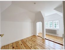 Ein heller, leerer Raum mit weiß gestrichenen Wänden und einem Holzdielenboden. Der Raum verfügt über mehrere Fenster mit Sprossen, die Tageslicht hereinlassen, sowie einen Heizkörper unter den Fenstern. Ein kleiner Torbogen trennt zwei Bereiche im Raum.