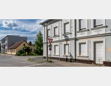 Straßenszene mit einem Eckgebäude aus hellem Putz mit geschlossenen Rollläden, daneben mehrere kleinere und größere Wohnhäuser, ein gelber Briefkasten und Verkehrszeichen an der Straßenecke. Der Boden ist mit Pflastersteinen bedeckt, vereinzelte Grünflächen sind zu sehen.