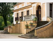 Historisches Gebäude mit creme-weißer Fassade und stuckverzierten Fensterelementen, flankiert von symmetrischen, geschwungenen Treppen aus Stein, die zu einem offenen Eingang führen. Der Vorbau ist mit dekorativen Blumenkübeln geschmückt, und im Hintergrund sind Bäume und eine grüne Landschaft zu sehen.