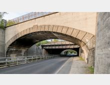 Straßenunterführung mit zwei übereinanderliegenden Brücken: eine massive Steinbogenbrücke im Vordergrund und eine Metallbrücke mit Geländern darüber. Darunter verläuft eine asphaltierte Straße mit Gehweg auf der rechten Seite.