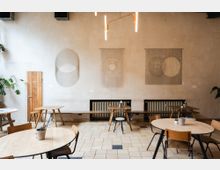 Ein minimalistisches Café mit hellem, schlichtem Interieur. Holzstühle und runde Tische stehen auf einem beige-karierten Fliesenboden; an der cremefarbenen Wand hängen abstrakte Kunstwerke, darunter Kreise und geometrische Formen.