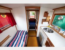 Innenraum einer kleinen Wohnkabine mit Holzrahmen. Links ein Einzelbett mit gestreifter Bettwäsche, rechts eine kleine Küchenzeile mit Spüle, Kühlschrank und Küchenutensilien; dahinter ein Durchgang mit Regalen und Zugang zu einer weiteren Schlafnische.