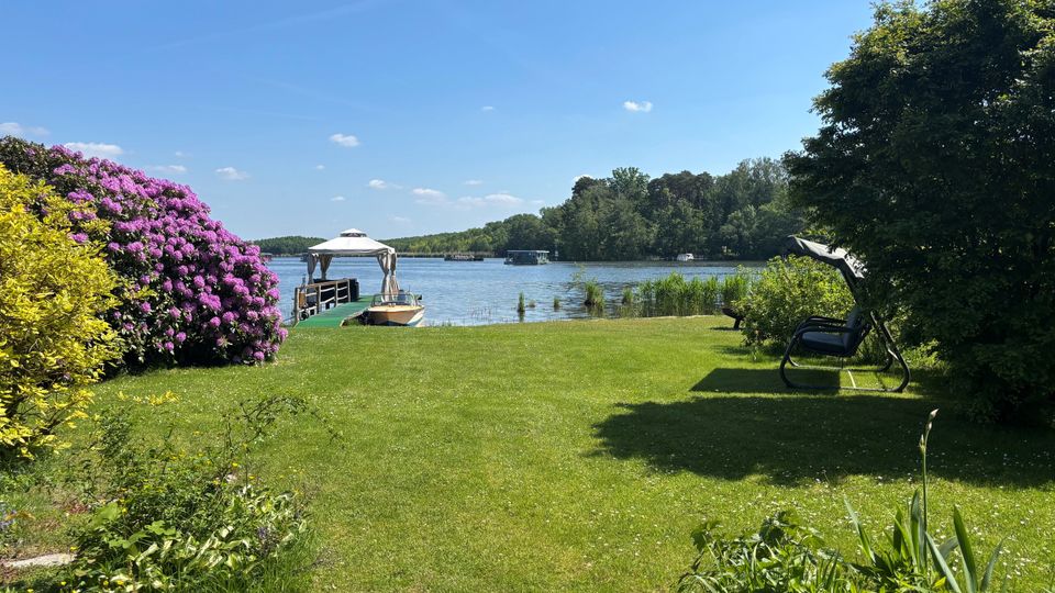 Grüne Rasenfläche am Ufer eines Sees mit Steg, kleinem Boot und weißer Überdachung. Links blühender Strauch, rechts Bäume und eine Gartenschaukel; gegenüber ein bewaldetes Ufer unter blauem Himmel.