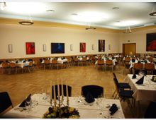 Ein Festsaal mit parkettiertem Boden, gedeckten Tischen mit weißen Tischdecken, blauen Servietten und Blumendekoration. An den Wänden hängen bunte, abstrakte Gemälde, und die Decke ist mit modernen Lampen beleuchtet.