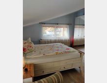 Schlafzimmer mit schräger Decke, einem Holzbett mit bunter Bettwäsche und einem Nachttisch mit Lampe. Ein Fenster mit weißen Vorhängen lässt Tageslicht herein, und ein großer Spiegelkleiderschrank reflektiert den Raum.