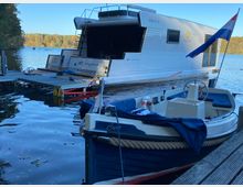 Ein modernes Hausboot mit der Aufschrift „The Kiss“ liegt an einem Steg auf einem ruhigen See, daneben ein kleines Motorboot mit blauen Sitzpolstern und einer niederländischen Flagge. Im Hintergrund sind Herbstbäume am Ufer sichtbar.